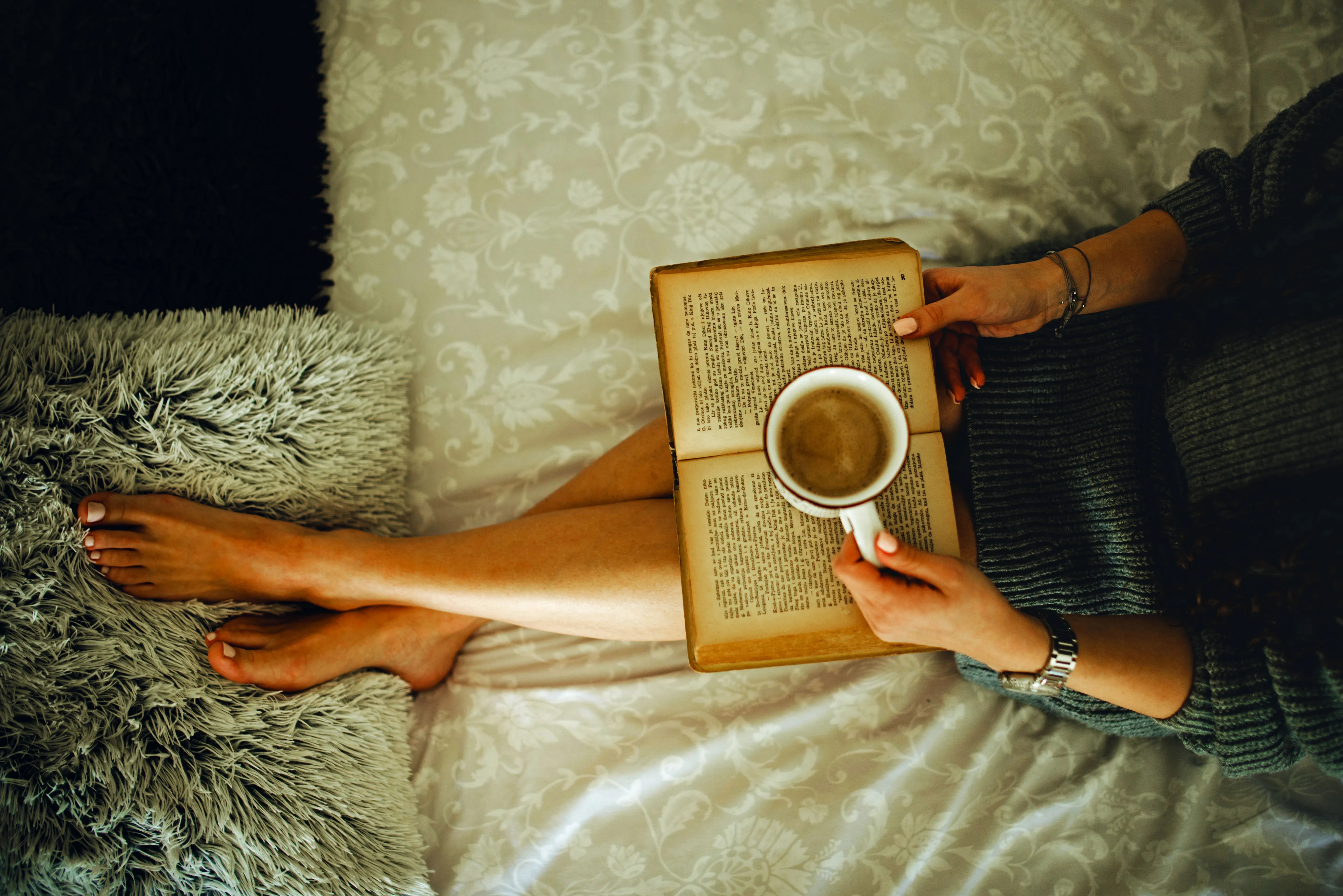 03 Woman sitting on her bed in a sweater while holding a cup of coffee and reading a book. 50894735221